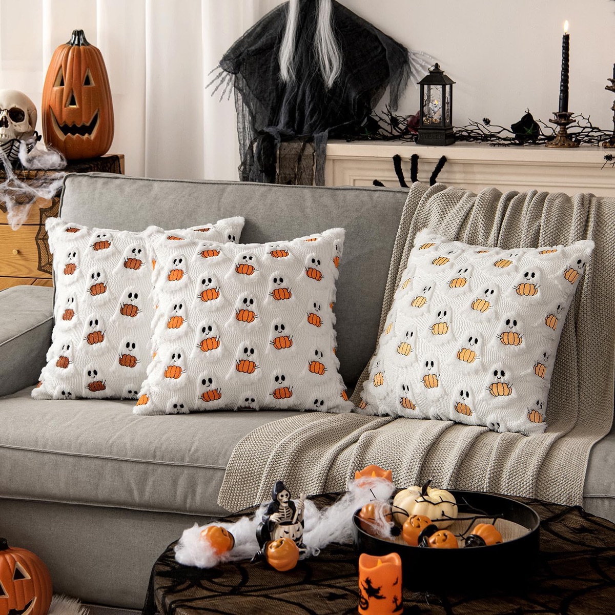 White plush pillows patterned with little ghosts holding orange pumpkins on a grey sofa with a knit throw