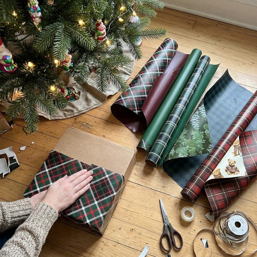 Hands wrapping a box in red and green plaid paper on a wooden floor beside the Christmas tree, with the four plaid and bear-print rolls unrolled nearby